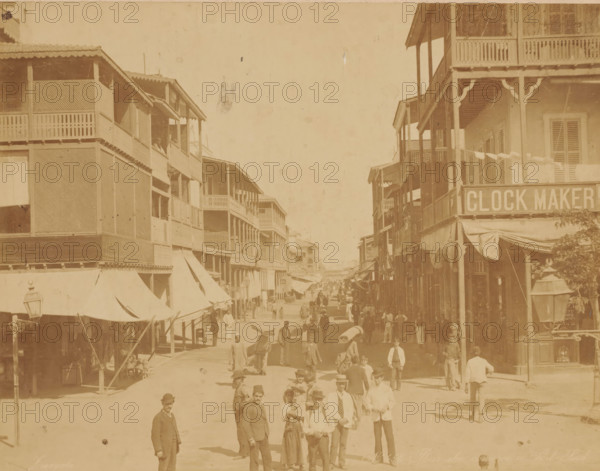 Shopping street in Port Said, Egypt, Rue du commerce a Port Said, photograph, number: 'No. 458', photographer: Zangaki, C. & G., Port Said, 1870 - 1910, North Africa, pasteboard, paper, albumen print, height 348 mm x width 445 mm, shop, store, street, A sunlit street scene lined with multi-story wooden buildings featuring balconies and awnings on both sides, with many people walking and standing along the road, some carrying bundles or wearing hats, and a lamp post visible in the foreground.