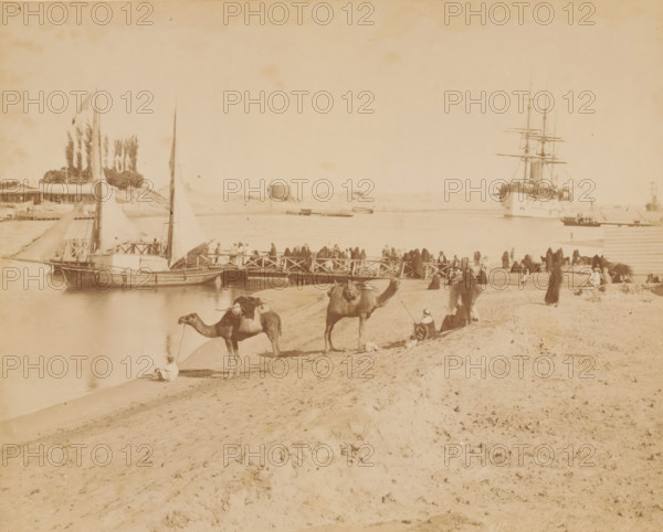 Pont de la Syrie à Kantara, Crossing point for caravans across the Suez Canal at Al-Qantarah, photograph, number: 'No. 110', photographer: attributed to Zangaki, C. & G., Al-Qantarah Gharb, 1870 - 1910, North Africa, Attributed to, pasteboard, paper, albumen print, albumendruk, height 219 mm x width 271 mm, height 348 mm x width 453 mm, canal, hoofed animals: camel, Suez Canal, A wide sandy shoreline with footprints and ridges extends across the foreground toward a busy riverbank where several camels stand or rest, some with saddles and packs, and nearby people sit or stand wearing flowing robes and head coverings; a wooden jetty projects into the water crowded with people, small sailboats moored alongside display tall triangular sails, and farther out a large multi-masted ship is anchored with rigging and spars visible against the sky, while low buildings, trees, and dunes line the opposite shore.