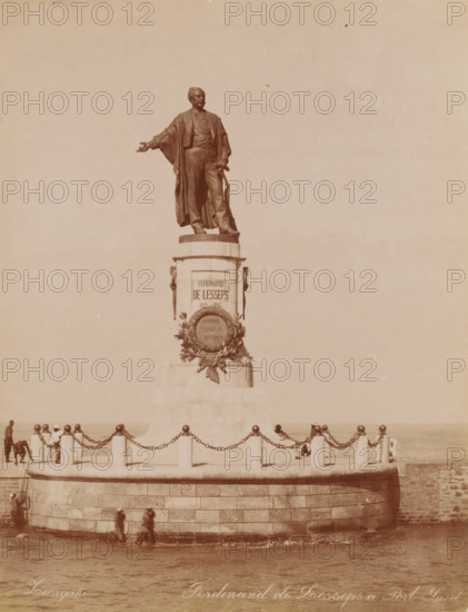 Statue of Ferdinand de Lesseps at the Suez Canal in Port Said, Ferdinand de Lesseps a Port Said, photograph, photographer: Zangaki, C. & G., Port Said, 1894 - 1910, North Africa, paper, albumen print, height 278 mm x width 215 mm, Person, Ferdinand de Lesseps, monument, statue, Suez Canal, A large bronze statue of a robed man stands atop a stone pedestal on a circular stone platform by the water, the figure shown with one arm extended and the other holding an object at his side; ornamental wreath or relief decoration is affixed to the front of the pedestal. The platform is surrounded by short stone bollards linked by heavy chains and a low curved stone wall, with a few people visible walking along the base of the platform near the water and a couple of figures near the bollards. The scene has a muted, sepia-toned appearance and an expanse of sky fills the upper portion of the image.