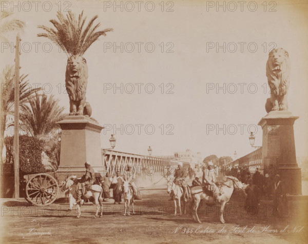 Entrance to the Qasr al-Nil Bridge, Qasr al-Nil Bridge over the Nile in Cairo, photograph, number: 'No 385', photographer: Zangaki, C. & G., Cairo (city), 1870 - 1910, Ottoman Empire, North Africa, paper, albumen print, height 223 mm x width 283 mm, bridge in city across river, canal, Qasr al-Nil Bridge, A wide sepia-toned scene shows a bridge flanked by two tall stone plinths each topped with a large lion statue, palm fronds rising behind the left lion and ornate street lamps mounted along the bridge. People and riders on donkeys and horses move across the bridge, some figures standing or walking, others seated on animals, and a wooden cart wheel is visible at the left foreground. Buildings and trees are visible in the distant background beyond the bridge, and the ground in the foreground appears dusty.