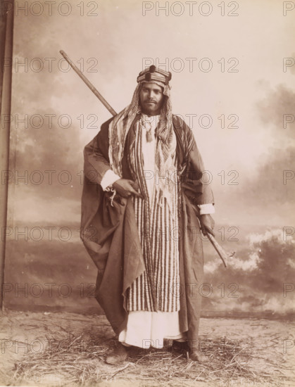 Costume Bedoin, Portrait of an unknown Bedouin man, photograph, number: 'N. 231', photographer: G. Lekegian & Co., Middle East, 1887 - 1900, paper, albumen print, height 212 mm x width 271 mm, anonymous historical person portrayed, wandering communities: nomads, A sepia-toned portrait of a man standing full-length against a painted backdrop showing clouds and distant landscape, wearing layered traditional garments including a long striped robe over a lighter ankle-length garment and a loose outer cloak, a patterned headscarf wrapped and secured with a band around his head, a tassel hanging at his chest, boots visible at his feet, a long staff resting diagonally over his shoulder, and a curved walking stick held in one hand, with straw scattered on the ground.
