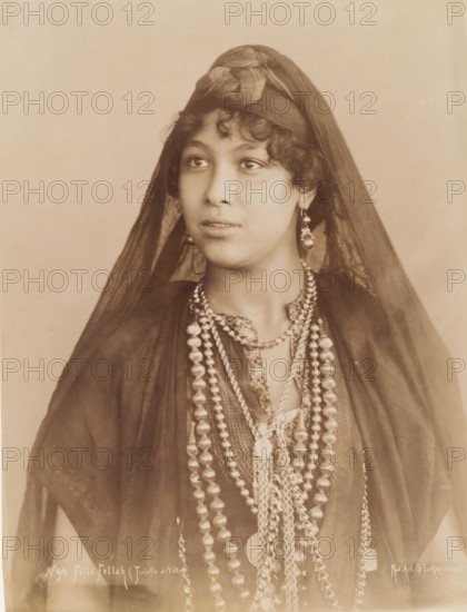 Fille Fellah (Toilette de Ville), Portrait of an unknown Fellah woman, photograph, number: 'No 94', photographer: G. Lekegian & Co., Middle East, 1887 - 1900, paper, albumen print, height 276 mm x width 212 mm, adult woman, anonymous historical person portrayed, A sepia-toned portrait of a young woman shown from the chest up wearing a dark sheer veil draped over her head and shoulders, a tied headscarf at the crown, curly hair visible at her forehead, large dangling earrings, multiple layered beaded necklaces of varying shapes and sizes, a patterned neckline beneath the necklaces, and a neutral plain background.