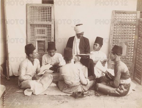 Ecole Arabe de jeuns garçons, School class with pupils and teacher, photograph, number: 'No 91', photographer: G. Lekegian & Co., Middle East, 1887 - 1900, paper, albumen print, height 210 mm x width 275 mm, teacher and pupil (pupil), A group of young men sit closely together on a tiled floor in front of patterned wooden screens, each holding open books and wearing long tunics and tall cylindrical hats, while a central figure with a wrapped headscarf and darker outer garment sits slightly elevated and looks down at an open book in his hands; footwear lies on the floor nearby and the scene is framed by carved lattice panels against a plain wall.