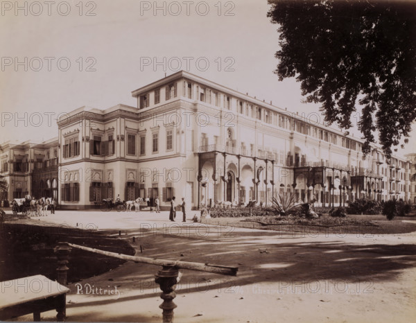 View of the Gezirah Palace Hotel in Cairo, Ghezireh Palace Hôtel, Cairo, photograph, number: '574', photographer: P. Dittrich, Gezirah, 1894 - 1900, Ottoman Empire, North Africa, paper, albumen print, height 210 mm x width 270 mm, hotel, hostelry, inn, Gezirah Palace, A large multi-story building with ornate façades and rows of shuttered windows occupies the center and right of the image, featuring decorative arches, columns, balconies with intricate railings, and an open arcade at ground level; horse-drawn carriages and horses stand in front of the building while groups of people in period clothing gather and walk near the entrance; a paved foreground with a low balustrade and ornamental posts leads toward a landscaped area with shrubs and designed plantings to the right; tall tree branches with dense foliage extend into the upper right corner, casting shadows across the scene.