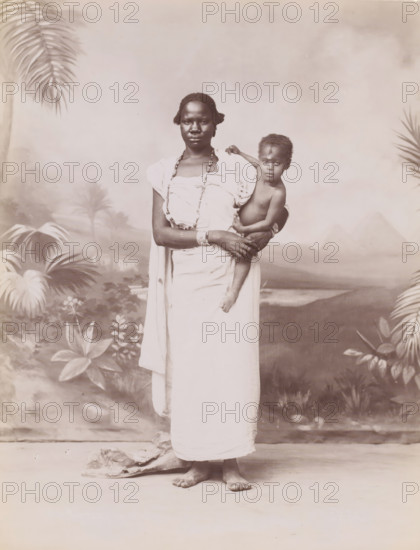 Portrait of an unknown African woman with child, Portrait of an unknown Nubian woman with child (former title), photograph, inscription: 'unreadable', photographer: Jean Pascal Sébah, Middle East, 1888 - 1900, paper, albumen print, albumendruk, height 270 mm x width 212 mm, anonymous historical person depicted, mother and baby or young child, Africans, A dark-skinned woman stands barefoot on a plain floor wearing a light cloth wrapped around her body and beaded necklaces, holding a dark-skinned child on her hip who is partially unclothed and looking toward the camera, with tropical plants and a painted backdrop showing palm trees and distant pyramidal shapes behind them.