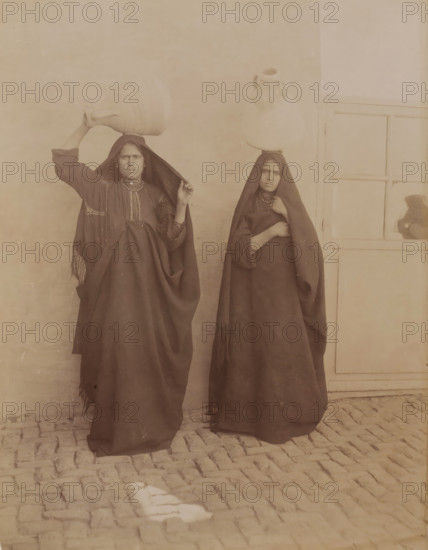 Double portrait of two unknown Egyptian women with jugs on their heads, Femmes Egypte Afalages ?, photograph, inscription: 'Dubbel', Middle East, 1860 - 1900, photographer, paper, albumen print, height 280 mm x width 219 mm, adult woman, Two women stand side by side against a plain wall and a panelled door, each wearing dark full-length garments with head coverings; the woman on the left holds a large vessel balanced on her head with one hand while lifting part of her head covering with the other, and the woman on the right also balances a vessel on her head while holding her garment closed with one hand; both wear necklaces and have neutral facial expressions, and cobblestones are visible beneath their feet.