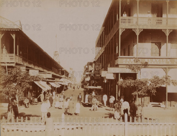 Rue du Commerce in Port Said, Rue du Commerce, Port Saïd, photograph, Port Said, 1860 - 1900, photographer, North Africa, paper, albumen print, height 215 mm x width 278 mm, street, A sepia-toned street scene shows two rows of tall wooden buildings with covered balconies and decorative railings flanking a wide avenue. People in period clothing walk and stand along the street and on the sidewalks, some grouped in small clusters and others moving toward the distance. A few horse-drawn carriages and a vehicle with an open cabin are visible among the pedestrians. Trees and awnings project from the buildings, casting shadows on the ground. Two figures stand on an upper balcony on the right building, and ornamental brackets accent the building corners. A low fence runs across the foreground.
