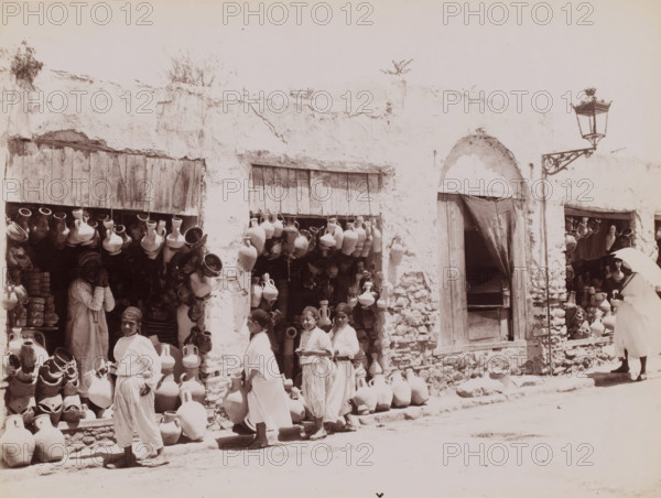 View of a jar shop, photograph, inscription: 'illegible', Middle East, 1860 - 1900, photographer, paper, albumen print, height 199 mm x width 257 mm, shop, store, container of ceramics: jar, jug, pot, vase, A sunlit street scene showing a worn white plaster building with wooden shutters and an arched doorway draped by a dark curtain, walls marked by cracks and patches of exposed stone; numerous clay jugs and pottery of various shapes hang from rafters, sit on shelves, and are stacked along the sidewalk and in shop openings; several people in light, flowing garments stand and smile near the pottery displays, including a group of young figures in the foreground and an adult partially visible inside a shop; a tall ornate street lamp on a decorative bracket projects from the wall near the arched doorway; the ground in front of the shops is dusty.