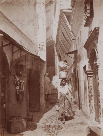 Unknown woman in an alley, possibly in Egypt, photograph, Egypt, 1860 - 1900, photographer, North Africa, paper, albumen print, height 246 mm x width 188 mm, adult woman, A narrow, sunlit alley flanked by tall, weathered buildings with textured walls and ornate doorframes, a woman in layered clothing and a headscarf walking toward the camera carrying a bundle on her head and holding a small tied sack, another figure partly visible further up the alley wearing a cloak, wooden awnings and beams projecting from the walls, a cobblestone path with uneven stones and shallow steps, hanging containers and tools affixed to the left wall, and shadows cast across the ground and lower portions of the façades.