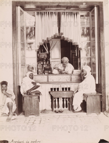 Perfume merchant, Perfume seller in his shop, presumably in Tunis, photograph, number: 'N. 44.', Tunis, 1860 - 1900, photographer, North Africa, paper, albumen print, height 259 mm x width 199 mm, shop, store, using scents, perfumes, ointments, A shopfront with an open window framed by wooden panels and a hanging vertical blind made of long thin rods, revealing an interior shelf filled with bottles and containers. A man leans on the counter inside, wearing a patterned headwrap and a loose garment, with his arms resting on the wooden ledge and a relaxed expression. Two other men sit on wooden stools outside the counter, both wrapped in light-colored draped robes and head coverings; one sits cross-legged with bare feet visible and an arm resting on his knee, the other sits facing slightly toward the counter with hands near his face. A small wooden table with a carved balustrade front stands between the stools, and a pair of sandals lie beneath it on the cobbled ground. The shop windows o...