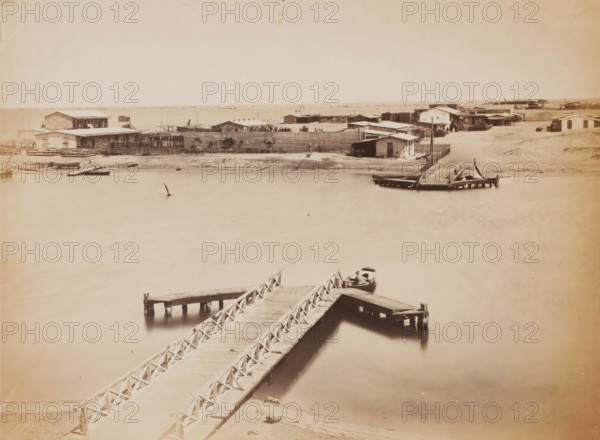View of a ferry, possibly in Egypt, photograph, Egypt, 1860 - 1900, photographer, North Africa, paper, albumen print, height 202 mm x width 270 mm, ferry, A sepia-toned waterfront scene showing a wooden pier with crisscross railings extending from the foreground into the water and branching into smaller platforms, a small boat moored alongside one platform, a blurred boat near the pier, calm reflective water filling much of the frame, a sandy shoreline across the water lined with low wooden buildings and fencing, several simple houses and sheds clustered behind the fence, additional docks and small boats along the far shore, and an expansive pale sky above.