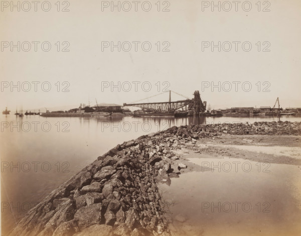 View of a harbor, photograph, 1860 - 1900, photographer, paper, albumen print, height 224 mm x width 288 mm, harbor, A wide view of a calm waterfront with a rocky breakwater leading from the foreground toward the middle distance, smooth water reflecting the shore, a shoreline across the water with boats moored and dock structures, a large metal crane-like structure and industrial equipment near the center, low buildings and piers lining the opposite shore, a few masts and smaller vessels visible near the docks, and a mostly open pale sky above.