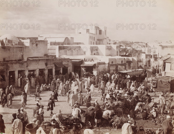 Mds de Légumes, Greengrocers on a square, probably in Tunis, photograph, number: 'No 84', photographer: J. André Garrigues, Tunis, 1860 - 1900, North Africa, paper, albumen print, albumendruk, height 199 mm x width 255 mm, market, A crowded outdoor market scene in front of a cluster of low, light-colored buildings with flat roofs and small windows, with many people wearing long robes and head coverings milling about, standing, walking, and sitting among baskets, bundles of produce, and market stalls, while some vendors display goods on the ground and others work around carts and awnings, and a larger multi-story white building rises behind the row of shops, with a wide open sky above.