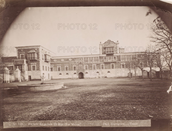 View of the Dar al-Taj in La Marsa, Façade du palais du Bey (la Marsa), photograph, number: 'N. 110.', photographer: J. André Garrigues, La Marsa, 1860 - 1900, North Africa, paper, albumen print, height 198 mm x width 258 mm, palace, Dar al-Taj, A wide sepia photograph of a large ornate building seen from an open courtyard, showing a long facade with decorative windows, balconies, and architectural details, with a central arched entrance and a raised ornate section above it, flanked by lower wings; a circular stone fountain basin sits in the foreground on bare ground, leafless trees line the right side near a wrought iron fence and gate, and a few small plants and architectural elements are visible near the left side of the building.