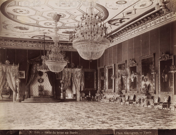 Salle du trône au Bardo, Throne Room in the Bardo Museum in Le Bardo near Tunis, photograph, number: 'N. 140.', photographer: J. André Garrigues, Le Bardo, 1860 - 1900, North Africa, paper, albumen print, height 200 mm x width 259 mm, throne-room, Bardo Museum, A large ornate interior with a high decorated ceiling featuring geometric and floral patterns and two massive crystal chandeliers suspended from it, an elevated throne area draped with heavy patterned curtains and framed by columns, richly upholstered chairs and sofas with carved wooden frames arranged along the right wall, a series of large framed portraits and mirrors with elaborate gilded frames hanging above side tables and candlesticks, patterned rugs covering the floor, decorative moldings and friezes along the upper walls, sculptural vases and ornamental objects placed on pedestals and tables, and overall lavish decorative detailing throughout the room.