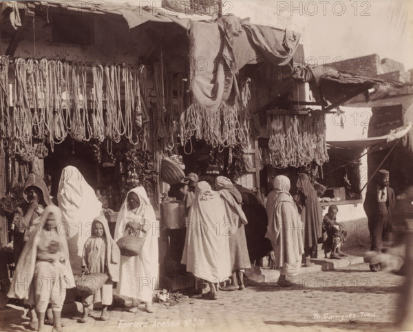 Grocery stores with customers, probably in Tunis, Epiciers Arabes, photograph, number: 'No 300', photographer: J. André Garrigues, Tunis, 1860 - 1900, North Africa, paper, albumen print, height 199 mm x width 248 mm, shop, store, A busy outdoor market scene showing a storefront with many ropes and bundles hanging from the awning and beams, fabrics draped over the top, and various goods displayed inside and below the hanging items; several people wearing long loose garments and head coverings stand and move in front of the shop, some carrying baskets or bundles, including adults and children; stone steps and a cobbled ground are visible beneath their feet; to the right a man wearing a cap stands near the building while a small child sits on a step holding an object; architectural elements of the shop and adjacent buildings, including rough walls and shaded recesses, appear in the background.