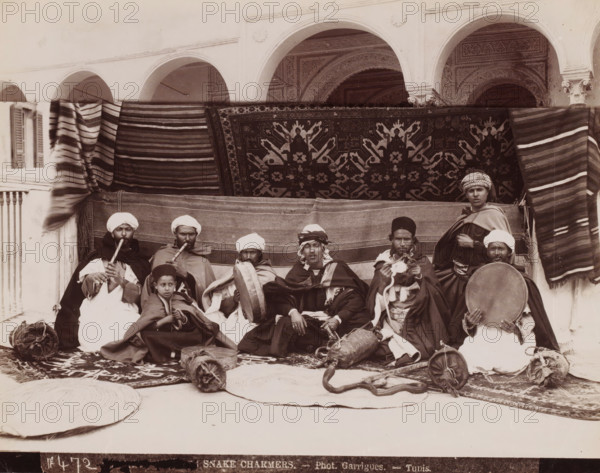 Snake Charmers, Snake Charmers in Tunis, photograph, number: 'No 472', photographer: J. André Garrigues, Tunis, 1860 - 1900, North Africa, paper, albumen print, height 199 mm x width 251 mm, snakes, A group of seated men and a boy arranged in front of patterned hanging rugs and striped cloths, many wearing turbans, caps, cloaks, and layered garments, with several holding or playing wind instruments and frame drums, one man holding a coiled snake on the ground near woven circular mats and bundled items, a background of arched architectural openings with decorative plasterwork and carved columns, and textile rolls and baskets placed around the group.