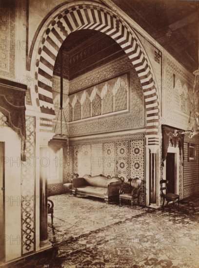 Interior of the Dar El Bey in the Medina of Tunis, Tunis, Dar-el-Bey, la Salle des Ministres, photograph, number: '367', photographer: Neurdein Frères, Medina of Tunis, 1863 - 1900, North Africa, paper, albumen print, height 273 mm x width 208 mm, palace, Dar El Bey, A richly decorated interior with a large striped archway featuring alternating light and dark bands framing a recessed area with ornate patterned walls and a carved frieze. Beneath the arch, tessellated wall panels display geometric and floral motifs. A carved wooden couch with upholstered seat and curved armrests sits against the patterned wall, flanked by matching upholstered chairs with carved legs and arms. Heavy draped curtains with fringed valances hang at openings, and a hanging chandelier with chains descends into the space. The floor is covered by an intricately patterned carpet extending across the room. Columns and pilasters with decorative inlays and carved capitals support the arch and frame doorways, and smal...
