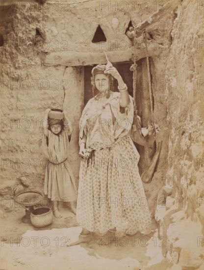 Unknown woman with spinning wheel, behind her a child, photograph, inscription: 'illegible', photographer: Neurdein Frères, Middle East, 1863 - 1900, paper, albumen print, height 275 mm x width 208 mm, adult woman, child, spindle, A sepia-toned photograph showing part of a rough stone or mud wall and a narrow doorway with a horizontal wooden beam above it, a woman standing in the doorway wearing a patterned skirt and loose top with layered fabric and bracelets on her wrists, holding a long narrow object vertically above her head with one hand while the other hand hangs by her side, and a child standing beside her with a cloth wrapped around their head and holding a small round basket or pot on top of their head, both barefoot on a worn earthen floor with two round vessels placed nearby and a hanging rope or strap suspended from the beam to the right.