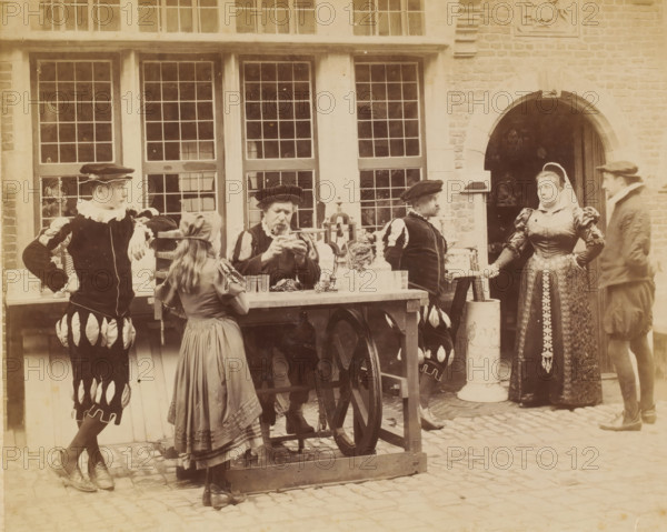 People in theatrical costumes for the occasion of the 1894 World's Fair in Antwerp, Belgium, Antwerp Exposition universelle 1894, photograph, check stamp: '...', number: '3', inscription: 'Tous droits réservés ...', photographer: AL, Antwerp, publisher: Edgard Lyon-Claesen, Brussels, 1894, AL, Edgard Lyon-Claesen, photographer, publisher, pasteboard, paper, albumen print, height 270 mm x width 352 mm, historical costume, Wereldtentoonstelling 1894 (Antwerp), Period, A group of people wearing historical-style clothing stand and sit in front of a building with tall multi-pane windows and a brick wall with an arched doorway; a wooden table on wheels holds glassware and decorative objects while a seated man at the table examines something in his hands, a young girl in a dress stands at the table facing him, and several other figures in puffed-sleeve garments and caps stand nearby with one woman in a patterned gown and head covering standing near the arched doorway.