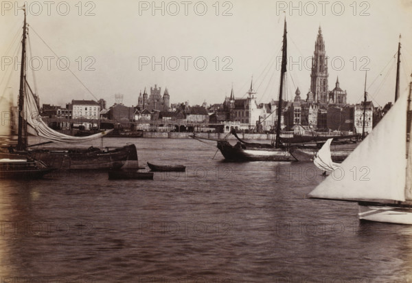 View of Antwerp from the Scheldt, Belgium, photograph, Antwerp, 1851 - 1900, photographer, paper, albumen print, height 190 mm x width 280 mm, sailing-ship, sailing-boat, city-view in general, 'veduta', parts of church exterior and annexes: tower, Scheldt, Cathedral of Our Lady (Antwerp), A waterfront scene showing several sailing vessels moored on calm water with visible hulls, masts, rigging, and partially furled sails, a small rowboat floating near the larger boats, and a large triangular sail in the foreground at the right; across the water a stone quay and a row of buildings rise with varied rooflines, towers, spires, and a prominent tall gothic-style tower dominating the skyline, all under a pale sky.