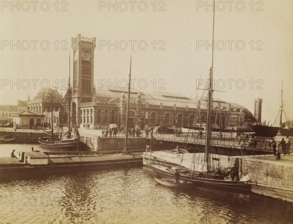 Ostende. La gare, View of the station of Ostend, Belgium, photograph, inscription: 'M&T', inscription: '... Anvers ?', number: 'No 74', Ostend, 1882 - 1900, photographer, paper, albumen print, albumendruk, height 215 mm x width 279 mm, station, shelter (railway, tramway), Ostend-City Station, A waterfront scene shows a large brick building with a tall clock tower and a long arched roof, bordered by a stone quay and a metal railing where people stand and walk; several masts, rigging, and small sailing vessels are moored along the quay, with crew or workers visible on and beside the boats, and the water in the foreground reflects ripples and shadows from the boats and structures.