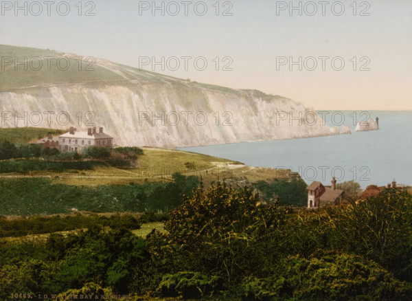 View of Alum Bay on the Isle of Wight, in the distance on the right the Needles, IOW Alum Bay & Needles, photomechanical print, number: '10469.', inscription: 'Knight's Library, 147 & 149, High St., and, Bank Buildings, Church St., Ventnor, IW', Alum Bay, cliché maker: Photochrom Zürich, Zürich (city), publisher: Photochrom Zürich, 1889 - c. 1920, Photochrom Zürich, photographer, cliché maker, publisher, Isle of Wight, paper, photochrom, height 165 mm x width 225 mm, coast, Needles, The, A coastline scene showing pale chalk cliffs rising above a calm blue sea, with a small cluster of jagged rock formations jutting from the water near the cliff edge and a dark vertical structure on one of the rocks. In front of the cliffs there are sloping grassy fields with a large pale building set back among low trees and hedges, and nearer to the foreground a smaller stone house with a pitched roof and chimney partially obscured by foliage. The foreground is filled with dense green bushes and trees,...