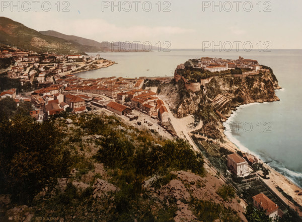 Monaco, View of Monaco, photomechanical print, number: '1082.', cliché maker: Photochrom Zürich, Zürich (city), 1889 - c. 1920, photographer, paper, photochrom, height 165 mm x width 223 mm, city-view in general, 'veduta', A coastal town seen from a high vantage point with a rocky cliffside jutting into a calm sea, a densely packed cluster of red-roofed buildings and narrow streets spreading across the slope, terraces and pathways carved into the cliff, a fortified promontory topped by larger structures and greenery, a harbor inlet with a few small boats on the water, rolling hills and more buildings in the background, vegetation and rocky ground in the foreground, and a pale sky above.