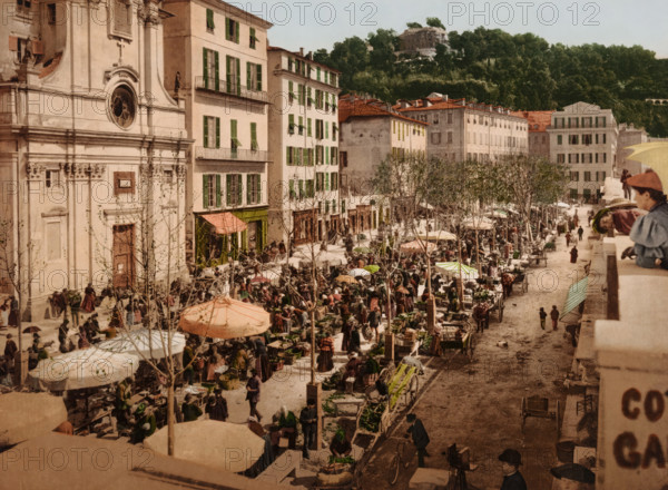 Nice, le Marché, View of the market in Nice, bottom right a photographer with camera, photomechanical print, number: '1759.', cliché maker: Photochrom Zürich, Zürich (city), 1889 - c. 1920, photographer, Provence-Alpes-Côte d'Azur, paper, photochrom, height 167 mm x width 226 mm, market, photographer at work, A wide street market scene with rows of stalls and umbrellas stretching along a tree-lined avenue beside tall pale buildings with green shutters and balconies, a large ornate building facade on the left with a round window and carved details, many people milling between carts and stalls carrying baskets and goods, vendors and shoppers gathered under patterned umbrellas and awnings, wooden carts and produce displayed on tables, leafless trees planted along the street, a hillside covered with dense trees and a stone structure visible above the treeline in the background, and a stone balustrade in the foreground where several children and a few adults lean and look down toward the ma...