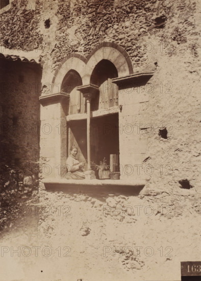 Unknown woman in a window, presumably in Italy, photograph, number: '163', photographer: Giovanni Crupi (possibly), Italy, 1865 - 1875, paper, albumen print, height 166 mm x width 120 mm, adult woman, open window, A stone building facade with rough textured walls and two arched window openings joined by a central column, a woman seated on the window ledge wearing a long skirt and blouse while holding an object, a wooden shutter or door behind her, a potted plant and a basket placed on the ledge beside her, scattered stones and rubble at the base of the wall, and small irregular holes in the masonry.
