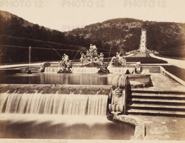 Fountain in the garden of the Palace of Caserta, Italy, Caserta Cascade Emblemes Sicile, photograph, number: 'No 4600', photographer: attributed to Giorgio Sommer, Caserta, 1857 - 1914, Attributed to, pasteboard, paper, albumen print, height 306 mm x width 403 mm, ornamental fountain, Palace of Caserta, A wide stone terrace with cascading water features fills the foreground, showing a stepped waterfall flowing into a rectangular pool with smooth, blurred water; ornate carved balustrades and stone steps run along the right edge, one carved face visible on a curved corner. In the middle distance a large sculptural fountain complex sits on a raised platform, with multiple statues of human figures and animals and jets of water arcing from several points into the basin below. Further back formal lawns and pathways extend toward a tall vertical water feature at the far end, bordered by trimmed hedges that create terraced levels. Behind the formal garden a densely wooded hillside rises across...