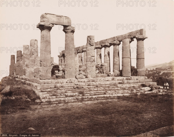 Ruin of the temple of Hera Lacinia in Agrigento, Girgenti. Tempio di Junone, photograph, number: 'No. 1833.', inscription: 'Rive (t)', photographer: attributed to Roberto Rive, Agrigento, 1860 - 1889, Sicily, Attributed to, pasteboard, paper, albumen print, height 197 mm x width 249 mm, height 307 mm x diameter 384 mm, ruin of a building, architecture, Temple of Hera Lacinia, A sepia-toned photograph of ancient stone ruins showing a row of tall, fluted columns standing on a stepped stone platform with some columns topped by large horizontal lintels and others partially broken, rough-hewn blocks and rubble around the base, sparse grass and low vegetation growing between the stone steps, an open sky above, and a person seated on a stone at the far right edge of the scene.