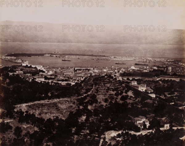 View of Messina, Italy, Messina Panorama, photograph, number: 'No. 1330', photographer: Giorgio Sommer, Messina, 1857 - 1914, Sicily, paper, albumen print, height 196 mm x width 240 mm, harbor, prospect of city, town panorama, silhouette of city, A wide sepia-toned panoramic view shows a coastal town and harbor seen from an elevated vantage point, with a foreground of terraced hillsides covered in trees and scattered houses, a midground of dense clustered buildings with varied rooflines and a few domed structures, and a background of calm water dotted with several ships and small boats alongside a low shoreline and distant hills under a pale sky.