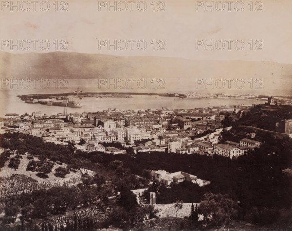 Panorama dal Casteluggio, Messina, View of Messina, Italy, photograph, inscription: 'Sommer', number: '8627', number: 'No. 1359', photographer: Giorgio Sommer, 1857 - 1914, Sicily, paper, albumen print, height 196 mm x width 241 mm, city-view in general, 'veduta', gulf, bay, A wide sepia-toned aerial view of a coastal town with densely packed buildings and tiled roofs in the foreground leading down to a harbor and a curved pier extending into calm water, with several ships anchored alongside the pier and a low mountain or hillside visible across the water in the background; scattered trees and cultivated plots appear on the slopes in the lower part of the image, and the sky above is pale and lightly mottled.