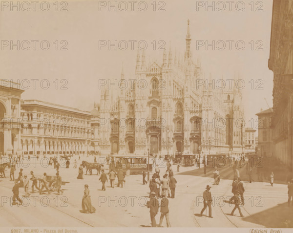 Piazza del Duomo, View of the square in front of the Milan Cathedral, Italy, Milano, photograph, number: '9987.', photographer: Carlo Brogi (possibly), Milan, publisher: Edizioni Brogi, Florence, 1881 - 1900, Tuscany, Lombardy, pasteboard, paper, albumen print, height 309 mm x width 399 mm, square, circus, church (exterior), Dom (Milan), A wide city square with a large ornate cathedral façade featuring tall spires and arched windows dominating the scene, flanked by multi-story classical buildings with rows of arches and columns, a busy street area in front with trams and horse-drawn vehicles, numerous pedestrians in period clothing walking or standing across the square, and people gathered near the tram and building entrances.