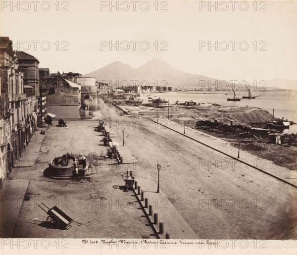 View of the harbor of Naples and Mount Vesuvius, Italy, Marine, Chateau Carmine-Vesuve avec Portici, Naples, photograph, number: '12 -', number: 'No 4008', number: '12', photographer: attributed to Giorgio Sommer, 1857 - 1914, Attributed to, pasteboard, paper, albumen print, height 307 mm x width 404 mm, harbour, volcano, A wide waterfront scene showing a broad paved promenade running alongside a shoreline with scattered debris and wooden planks, a row of short bollards lining the edge, and several lampposts along the promenade; to the left there are multi-story buildings with balconies and arched doorways, a circular stone fountain with figures, and a few handcarts and wagons positioned on the pavement; in the middle distance there are low stone structures and small waterfront platforms and docks extending into the water; on the water two sailing vessels with tall masts are anchored near the shore; in the background a large double-peaked mountain rises with a faint plume of smoke or h...