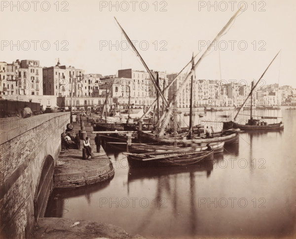 Harbour view in Naples, Italy, Naples la Marine, photograph, number: '12', number: '13.', Naples, 1851 - 1900, photographer, pasteboard, paper, albumen print, albumendruk, height 306 mm x width 404 mm, harbour, sailing-ship, sailing boat, A waterfront scene showing several wooden sailboats moored along a stone quay with tall, angled masts and rigging rising above the boats, calm reflective water, a stone wall and stepped edge along the quay with bollards and coiled ropes, a few people standing and sitting on the quay near the boats, and a long row of multi-story buildings with balconies and windows lining the opposite shore.