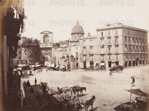 View of a square with the Porta Capuana and the dome of Santa Caterina a Formiello in Naples, Italy, Port Capuane, Naples, photograph, number: '17', number: 'No. 4067', number: '12', photographer: attributed to Giorgio Sommer, 1857 - 1914, Attributed to, pasteboard, paper, albumen print, albumendruk, height 306 mm x width 403 mm, square, circus, city-gate, parts of church exterior and annexes: dome, Porta Capuana, Santa Caterina a Formiello, A wide sepia street scene showing a large open plaza bordered by multi-story stone buildings with rows of shuttered windows and small balconies, a domed church rising behind the buildings, an ornate archway set into an older stone structure, and smaller storefronts and awnings along the base of the buildings; in the plaza there are horse-drawn carts and wagons, several groups of people standing or walking, market stalls with canvas coverings, barrels loaded on a wagon, and a few figures sheltered under an umbrella near the foreground, while the lef...