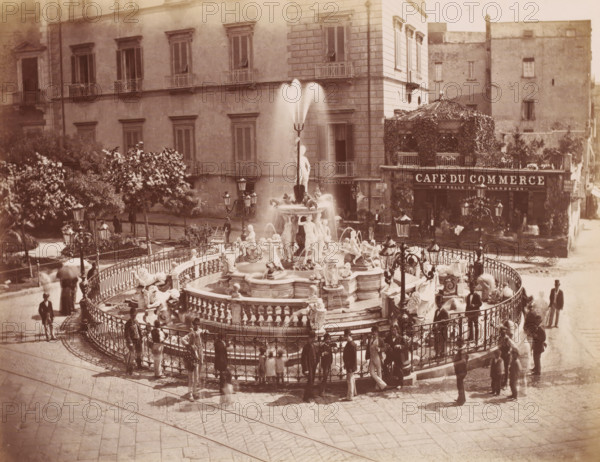 Naples fontaine Médine, Neptune fountain in Naples, Italy, photograph, number: '12', number: '21.', Naples, after sculpture by various manufacturers, 1851 - 1900, various manufacturers, photographer, after sculpture by, pasteboard, paper, albumen print, height 307 mm x width 403 mm, ornamental fountain, A large ornate fountain with sculpted figures and water jets stands in the center, surrounded by a decorative iron railing and several lamp posts; people in period clothing gather around the railing and walk nearby, some carrying umbrellas and some wearing hats; behind the fountain is a multi-story building with shuttered windows and small balconies, and to the side a façade covered in foliage with an awning and an entrance; trees and planted greenery are visible along the street, and the scene is set on a paved open square with stone paving patterns.