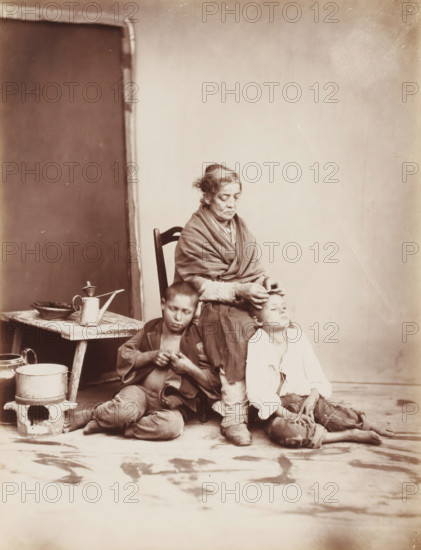 Naples, Scene in Naples with a woman removing fleas from the hair of two boys, photograph, inscription: 'Sommer (t)', photographer: Giorgio Sommer, 1870, pasteboard, paper, albumen print, height 384 mm x width 310 mm, looking for fleas - AA - female human figure, An older woman sits on a wooden chair wrapped in a shawl, her hair pulled back, with a solemn facial expression as she uses her hands near the head of a boy seated on the floor beside her; another boy sits nearby on the floor with a downcast gaze and hands held together, barefoot and wearing worn clothing. To the left, a small rustic table holds a watering can and a shallow bowl, and a low metal cooking pot sits on a small stand on the floor. The background shows a plain wall with a darker rectangular panel at one side, and the floor in front has visible scuffs and marks.