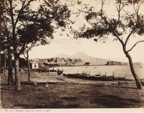 View of Naples, Castel dell'Ovo and Vesuvius, Italy, Prize du Jardin royal, Naples, photograph, number: '12', number: '10-', number: 'No. 4051', photographer: attributed to Giorgio Sommer, 1857 - 1914, Giorgio Sommer, photographer, Attributed to, pasteboard, paper, albumen print, height 305 mm x width 403 mm, gulf, bay, landscape with tower or castle, volcano, Castel dell'Ovo, Mount Vesuvius, A sepia-toned coastal scene framed by tall trees with leafy branches in the foreground, a shoreline with small wooden boats pulled up on the beach and lined along the water, a low stone wall and a small building near the edge of the shore, a calm bay with a few boats floating, a densely built hillside town across the water with clustered buildings of varying heights, and a large gently sloping mountain rising behind the town, all under a pale sky.