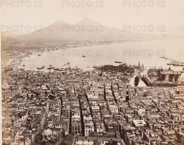 View over the Bay of Naples and Mount Vesuvius from the (former) Carthusian Monastery of Saint Martin, Italy, Panorama prize de la Chârtreuse S. Martin, Naples, photograph, number: '12', number: 'No 4003', number: '1-', photographer: attributed to Giorgio Sommer, 1857 - 1914, Attributed to, pasteboard, paper, albumen print, height 306 mm x width 405 mm, city-view in general, 'veduta', gulf, bay, volcano, Mount Vesuvius, Carthusian Monastery San Martino, A wide aerial view of a coastal city showing densely packed buildings with narrow streets running toward a waterfront lined with boats and docks, a harbor with many small and larger sailing vessels anchored and a pier extending into calm water, a prominent fortress-like structure near the water with towers and walls, a lighthouse-like tower by the docks, a distant shoreline curving around the bay, and a large mountain with a twin peak rising beyond the city under a pale sky.