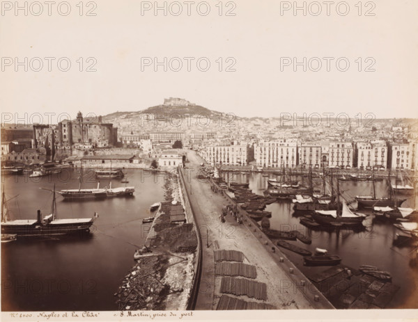 View of Naples and the Castel Sant'Elmo and the (former) Carthusian Monastery of Saint Martin, Italy, Naples et la Chartreuse S. Martin, prize du port, photograph, number: '12', number: '6-', number: 'No 4000', photographer: attributed to Giorgio Sommer, Naples, 1857 - 1914, Giorgio Sommer, photographer, Attributed to, pasteboard, paper, albumen print, height 307 mm x width 404 mm, harbour, city-view in general, 'veduta', landscape with tower or castle, Castel Sant'Elmo, Carthusian Monastery San Martino, A wide view of a harbor scene with a long stone quay running from the foreground toward the middle distance, small boats moored along both sides of the quay, and larger sailing vessels with masts and furled sails in the water on either side; a paved road on the quay with a few horse-drawn carts and people, stacks of long bundled materials laid out near the foreground, and stone bollards along the quay edge; on the left a large fortified stone structure with worn walls and a tower adjac...