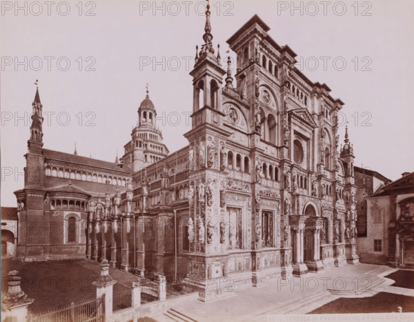 Exterior of the monastery church Certosa di Pavia in Pavia, Italy, Facciata e Fianco della Chiesa, Certosa di Pavia, photograph, number: '13', number: '4615.', inscription: 'Brogi (+)', Pavia (city), publisher: attributed to Giacomo Brogi, Florence, 1856 - 1881, photographer, Tuscany, Lombardy, Attributed to, pasteboard, paper, albumen print, height 306 mm x width 404 mm, church (exterior), A large ornate stone building fills the image, showing an elaborately decorated facade with carved statues set into niches, arched doorways, roundels, and layered architectural detailing; multiple towers and spires rise above the structure, including a domed tower with stacked cylindrical sections and a tall slender spire with finials; the lower foreground shows steps leading up to the entrance and a paved area bordered by a low fence with decorative posts; to the left and right are adjoining sections of brick construction with rows of arched windows and rooflines, and small sculptural elements and ...