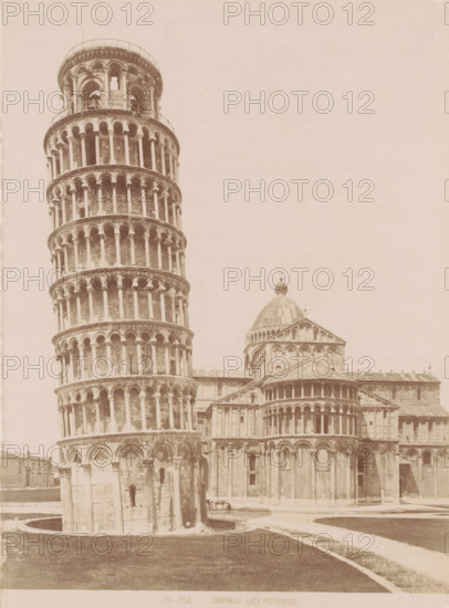 Tower of Pisa, Italy, Pisa Campanile Lato Posteriore, Pisa, photograph, number: '1004.', 1851 - 1900, photographer, Tuscany, pasteboard, paper, albumen print, albumendruk, height 385 mm x width 310 mm, parts of church exterior and annexes: campanile, A tall cylindrical stone tower with multiple tiers of open arches and slender columns leans noticeably to one side, standing in front of a larger stone building that features a dome with a decorative finial and a triangular pediment above a row of arches; the foreground shows a grassy area and curving paved paths, and portions of the adjacent buildings and architectural details such as rounded arcades, pilasters, and carved stonework are visible.