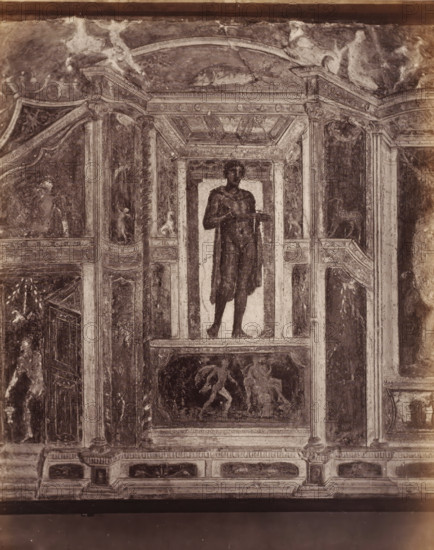 Photo reproduction of a fresco depicting a standing man in Pompeii, Italy, photograph, reproduction, number: '74.', inscription: 'Grand ... Pompei', number: '11', Pompeii, after painting by anonymous, 1851 - 1900, photographer, pasteboard, paper, albumen print, height 403 mm x width 307 mm, picture, painting, wall-covering, A faded wall painting framed by architectural painted columns and panels shows a full-length figure standing on a shallow platform within a recessed niche, the figure draped in a dark cloak and holding a shallow dish at chest height; above the niche a coffered painted ceiling and arch are visible, and below the niche a rectangular panel contains a scene of three partially rendered figures in motion; surrounding panels feature decorative motifs, small human and animal figures, fluted pilasters, and ornamental borders, all rendered in worn tones and exhibiting cracks and areas of surface loss.