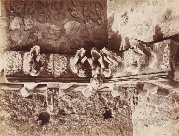 Decorated drain pipes in Pompeii, Italy, photograph, number: '11', inscription: 'Pompei, del gouttière, terre vite ?', number: '60.', Pompeii, 1851 - 1900, photographer, pasteboard, paper, albumen print, albumendruk, height 307 mm x width 405 mm, water supply and drainage, pipework (in house or building), A sepia-toned photograph showing a carved stone architectural element mounted on a rough stone wall, featuring a horizontal ornamental frieze with sculpted figures and decorative scrollwork; three reclining or leaning humanlike figures with detailed hair and drapery extend from the frieze, their hands raised and heads turned, and flanking them are carved animal-like or grotesque heads integrated into the ornamentation; beneath the frieze hang two ornate, symmetrical brackets with curled leaf motifs, and the surrounding wall displays irregular stone blocks and patches of lighter plaster or weathered surface.