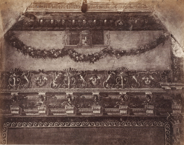 Photo reproduction of a fresco in Pompeii, Italy, photograph, reproduction, number: '70.', number: '11', Pompeii, after painting by anonymous, 1851 - 1900, photographer, pasteboard, paper, albumen print, height 304 mm x width 402 mm, picture, painting, wall covering, A section of an ornate wall or frieze with layered horizontal bands of decorative motifs, including a top cornice with a patterned border and a central recessed panel featuring a carved face peering from an opening, a swag of foliage and flowers draped below it, a middle register with scrolling acanthus designs, stylized vases, and human faces set among intricate curving ornament, and a lower register showing a row of egg-and-dart molding above a band of repeating spiral motifs and a darker field beneath; the surface appears aged and worn with visible cracks, spots of discoloration, and areas where the plaster or paint has chipped away.