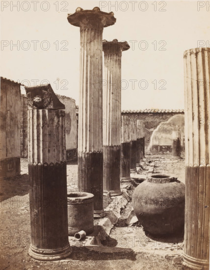 Remains of the house of Meleager in Pompeii, Italy, photograph, inscription: 'Pompei, maison Meleagre', number: '48', number: '11', Pompeii, 1851 - 1900, photographer, pasteboard, paper, albumen print, albumendruk, height 403 mm x width 305 mm, ruin of a dwelling, house, castle, A sepia-toned photograph showing a row of tall fluted stone columns with ornate capitals and weathered surfaces, some standing intact and others broken or shortened, set among low stone bases and fragments on the ground; a large rounded stone jar sits beside one column in the foreground, and a low basin and cylindrical stone drums are visible nearby; in the background there are worn plaster walls with a tiled roofline and patches of exposed masonry, and the ground is uneven with rubble and shallow trenches between the columns.