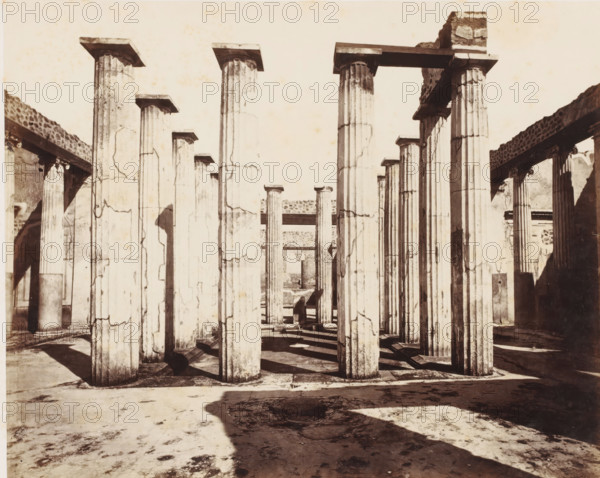 Remains of the house of Mario Epidio Sabini in Pompeii, Italy, photograph, number: '11', inscription: 'Pompei, maison Mario Epidio Sabini', number: '50', Pompeii, 1851 - 1900, photographer, pasteboard, paper, albumen print, albumendruk, height 305 mm x width 401 mm, ruin of a dwelling, house, castle, A monochrome photograph showing a row of tall fluted stone columns with square capitals standing on a paved floor, casting long shadows across the ground; some columns show surface cracks and weathering, and horizontal lintels span between a few of them; partial walls and additional columns are visible at the sides and in the background, with areas of rough stonework and shadowed recesses.